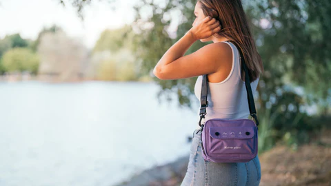 A brown haired woman stands facing a body of water. She is wearing a white tank top and jeans. She carries Sherpani crossbody, the Osaka in Iris, from the Open Your Eyes collection.