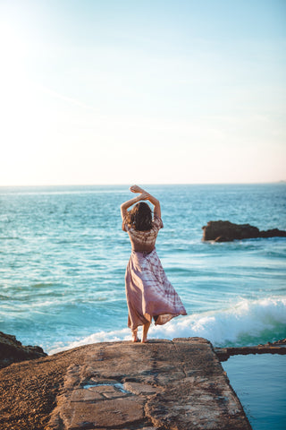 A dark haired woman stands on a rock racing the sea.