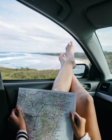 A woman sits in her car with a road map unfolded in her lap, her legs extending out the open window