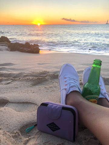 Sunset view from Maho Beach. On the sand, is a woman's feet, next to a Caribbean beer and her Sherpani RFID wallet, the Barcelona (Lavender color)