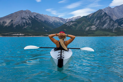 A blonde woman rests with her hands behind her head. She is alone in a canoe in blue water surrounded by mountaints.