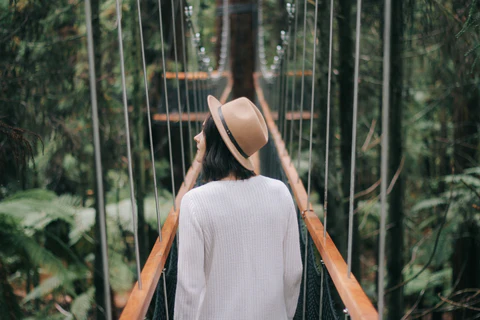 A woman stands on a bridge in a forrest, she is facing away from the camera and looking over her left shoulder.