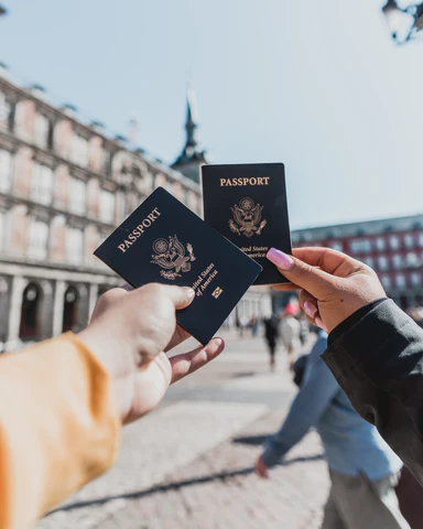 Two women hold out their passports in a scenic courtyard.