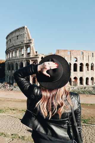 A blonde woman wearing a black hat faces away from the camera, in front of her is the Colosseum