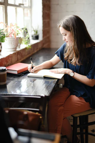 A woman sits at a desk in front of a window writing in her notebook.