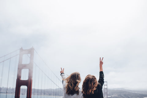 Two women stand side-by-side looking out at the Golden Gate Bridge making peace sign gestures.