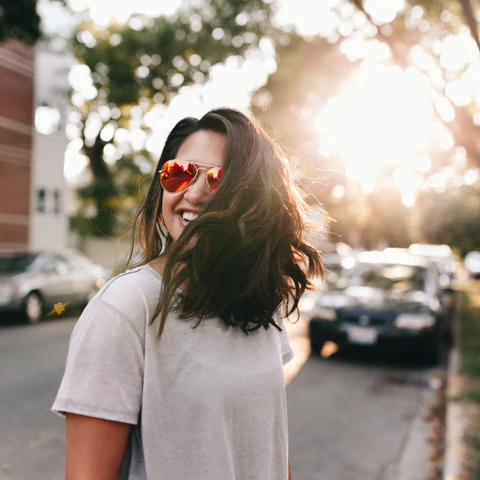 A brown-haired woman smiling to the distance. She is wearing a white tee shirt and sunglasses.