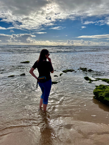 A woman standing in the ocean facing the horizon. She is wearing a black shirt, jeans and Sherpani's fanny pack the Hyk in Chromatic.