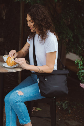 A woman eating a croissant at an outdoor table, she is wearing Sherpani Anti-Theft crossbody bag, the Vale