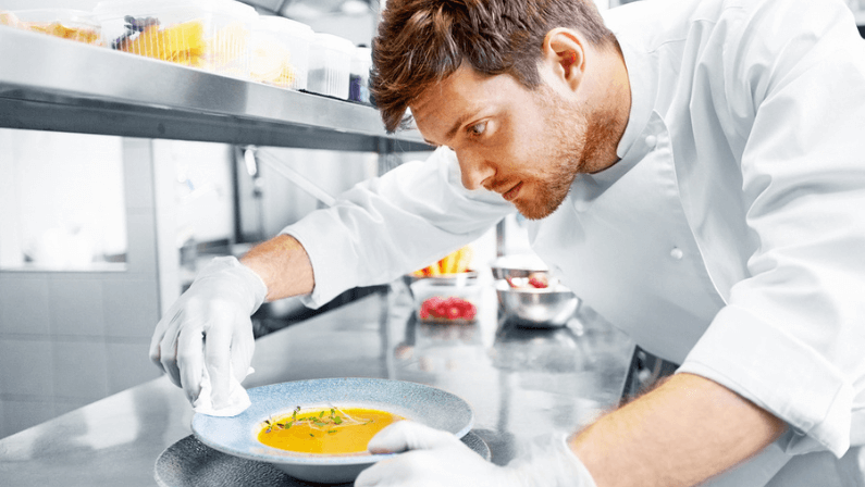 food cooking, profession and people concept - happy male chef cook serving and cleaning plate of soup at restaurant kitchen