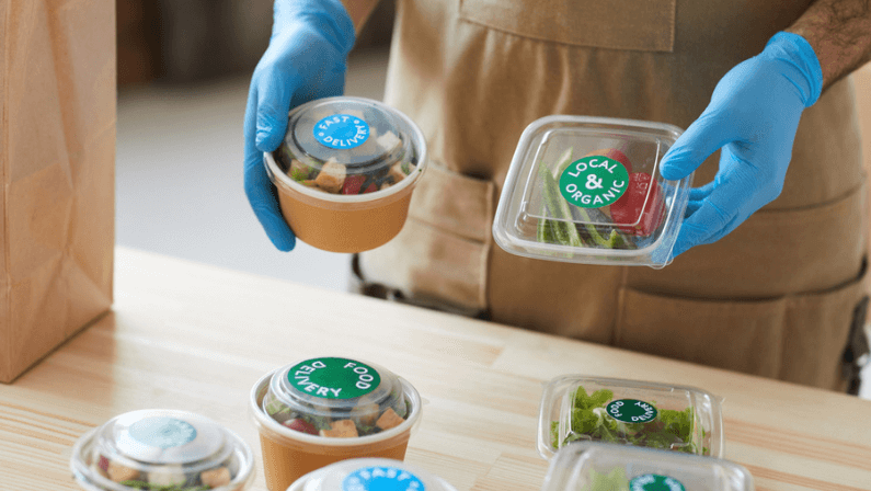 Closeup of unrecognizable worker wearing protective gloves safely packaging orders at wooden table in food delivery service, copy space