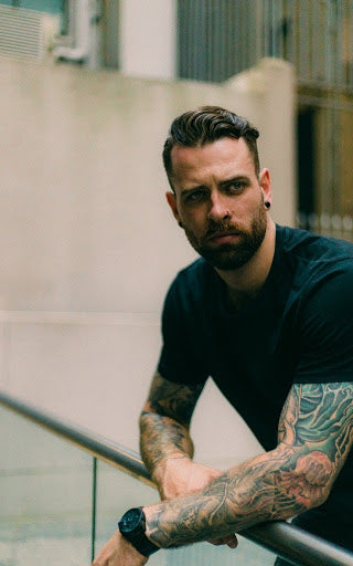 Man in t-shirt with tattoo sleeve leaning over a glass barrier | Style Standard