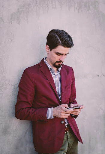 Man with goatee in red blazer, green chinos, and light striped shirt leaning against a wall and checking his phone | Style Standard