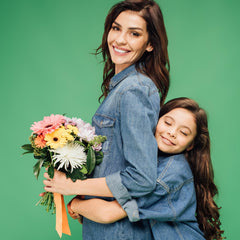 Unique family jewelry gifts for Mothers Day 2022 from RiseShed. A girl hugs her mother from behind. The mother is holding a bouquet of flowers in front of her. Both are facing to the left but looking towards the camera. Both mother and daughter are brunettes and wearing blue denim shirts.