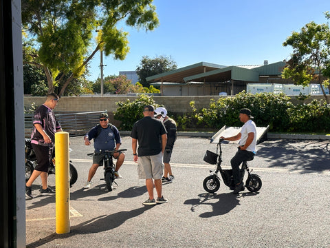 Men standing next to and sitting on electric scooters outside VoroMotors Kapolei store