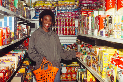 Image shows a woman in a sweatshirt holding a basket and standing in front of grocery store shelves filled with convenience food items.