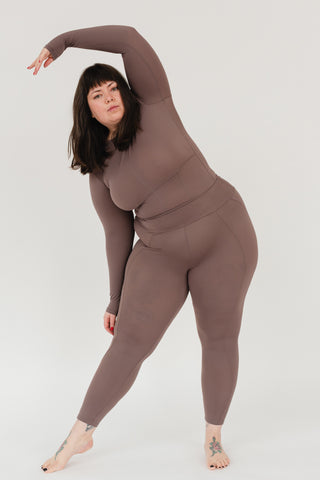 Image shows a woman wearing an all-brown unitard while standing in front of a white background. One arm is reaching above her head and she leans to one side in a gentle stretch.