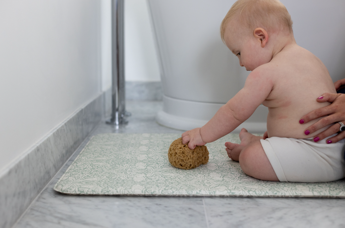 baby sitting on squishy runner in bathroom playing with natural sponge