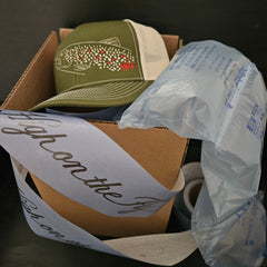 brown trout rising fly fishing hat sitting in box with packaging pillows and tape with high on the fly logo