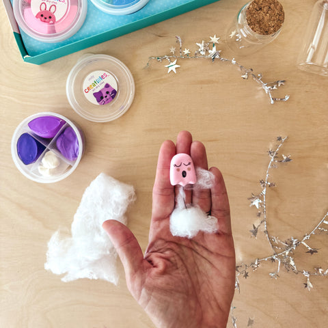 A hand holding a pink OOLY Creatibles air dry clay ghost with a small tuft of cotton stuffing, ready to be placed inside a glass bottle along with other decorative elements. OOLY craft Step 5.