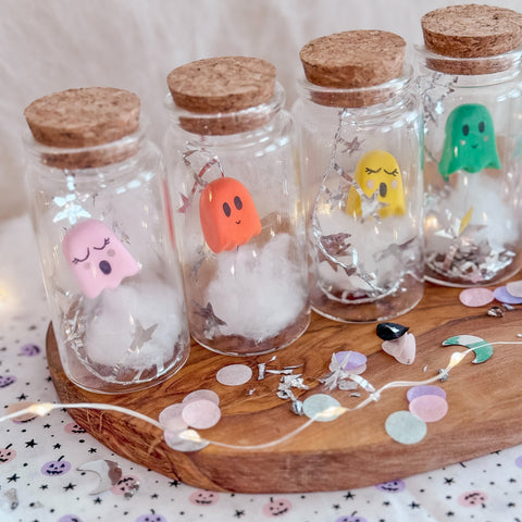 Four glass bottles with cork lids, each containing a brightly colored ghost made from OOLY Creatibles air dry clay, floating above cotton stuffing with star-shaped garland inside, placed on a decorative tray. OOLY Craft blog step 8.