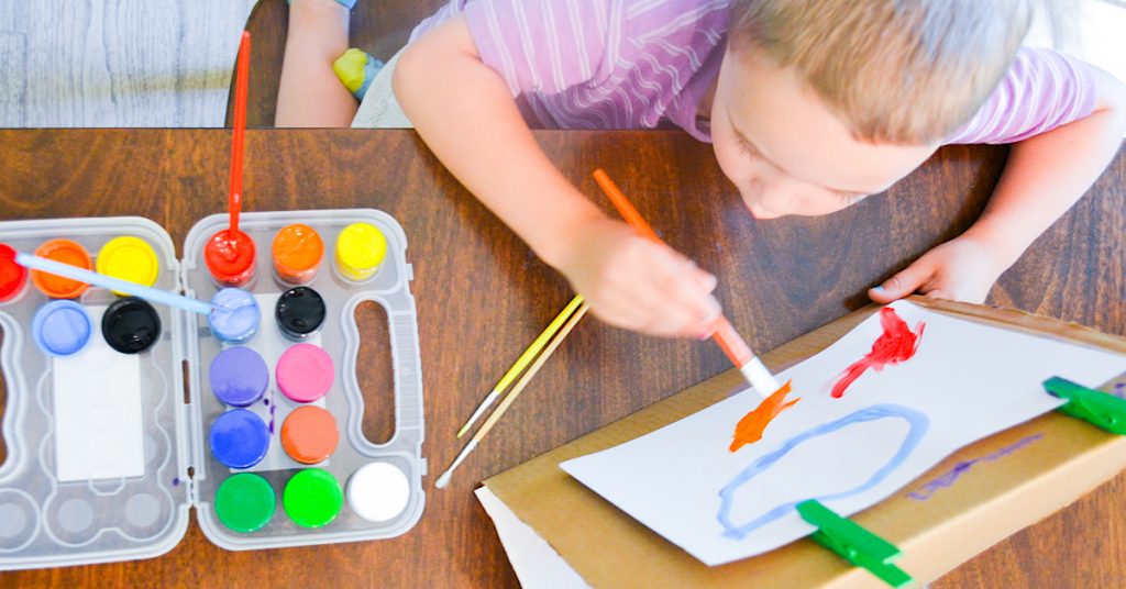 child in purple shirt sitting at table painting with orange brush