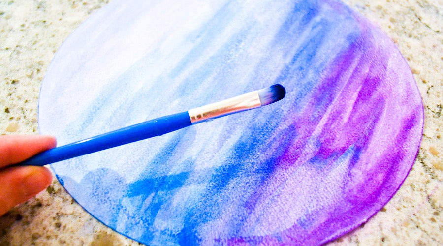 blue paint brush held in front of blue and purple watercolor on white circular paper