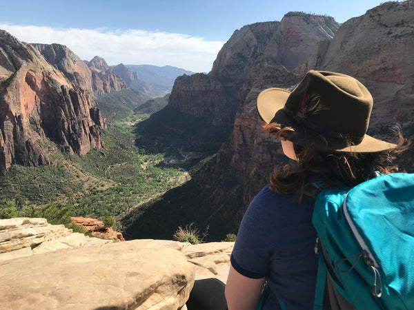 A woman in a hat sitting at the summit of Angel's Landing looking out into the valley of Zion National Park.