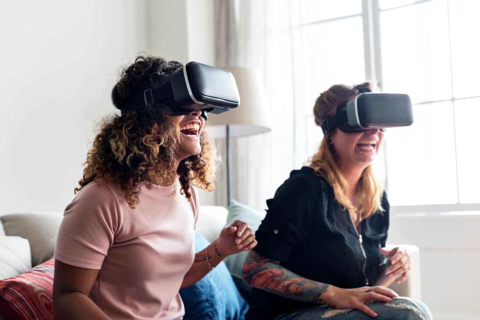 Two young women sit next to each other on a couch, both wearing virtual reality (VR) headsets. Both are laughing.