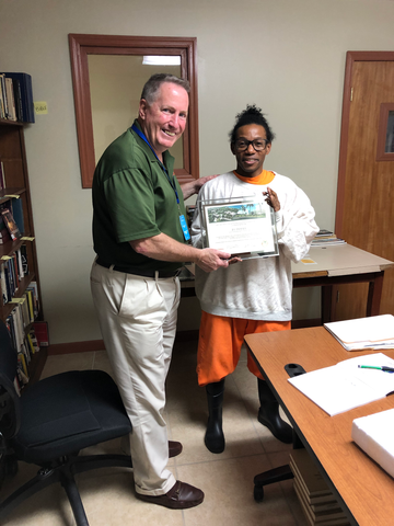 miek caslin presents gift certificate to man in orange jumpsuit and white sweatshirt