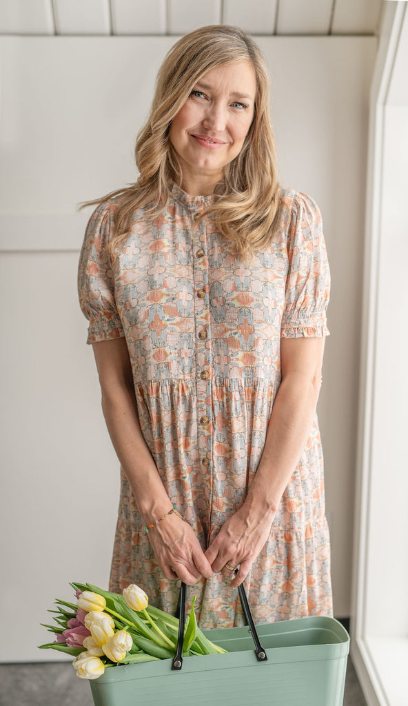 Dayna Manning wears a simple dress and smiles at the camera. She holds a basket containing tulips