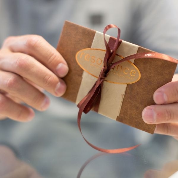 A man holds a decorated envelope. It is wrapped in brown and bronze paper and has bronze ribbon.