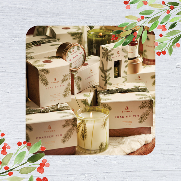 Thymes Frasier Fir votive candle is shown in the foreground. Behind it, boxes decorated in ivory paper with a Frasier Fir design on it are shown.