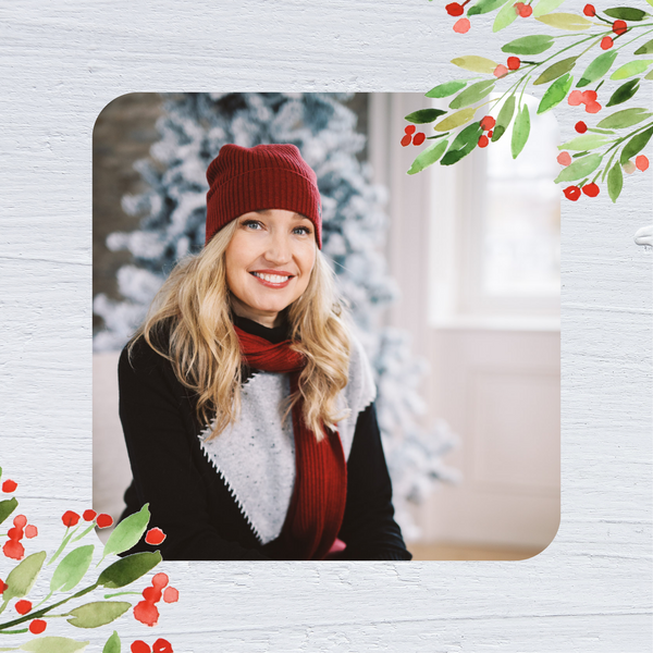 Dayna Manning is shown wearing  a Zaket & Plover sweater with a red scarf and hat. in the background, there is a snow encrusted holiday tree.