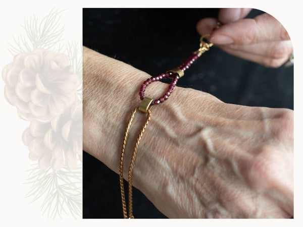 A woman holds a bracelet on her arm. It is made of copper coloured metal and small red stone beads.