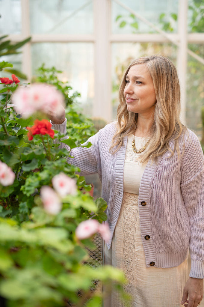 Dayna looks at some blooms in a greenhouse. She wears a lilac cardigan over an boho dress
