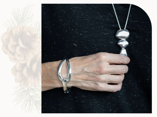 Silver jewellery is shown. A silver clasp bracelet in hammered metal is shown. The woman who wears the bracelet touches a silver pendant with three irregular silver balls in descending size.