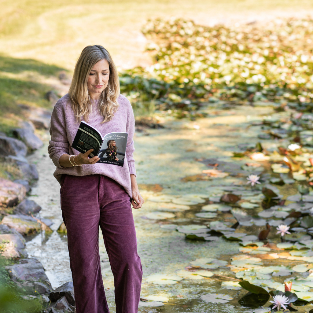 Dayna wears plum coloured cords and a dusty pink sweater as she walks along a river. She's holding a book.