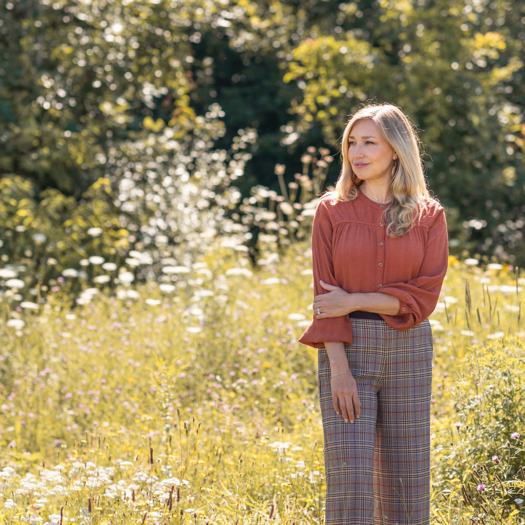 Dayna wears the same rust coloured blouse and checked pants, which have wide legs. She stands against a field of grasses and looks to one side.