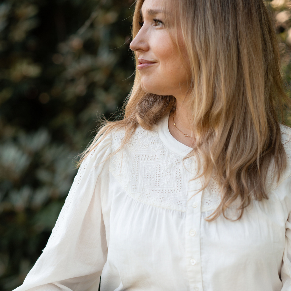 Embroidery is shown on the chest of Dayna's blouse, and down one sleeve. Dayna looks to the side and smiles slightly.