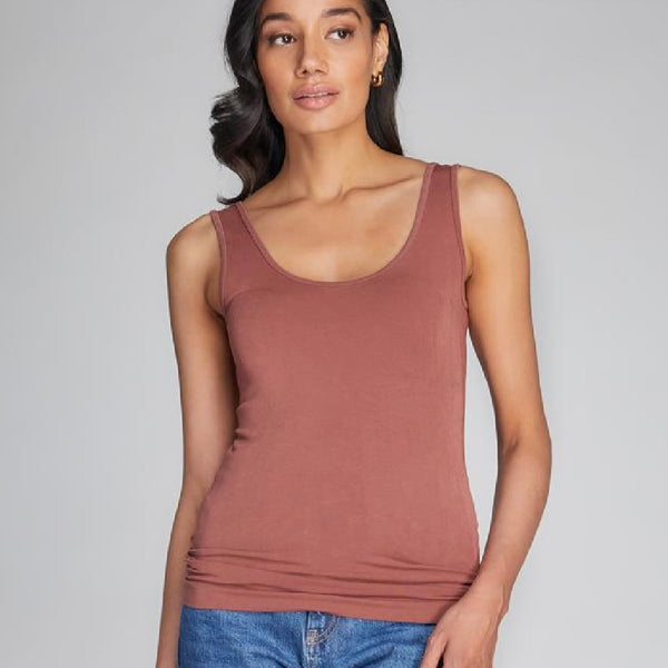 A woman wears a terracotta coloured tank top. It is long in the body and has thick shoulder straps. It is very fitted.
