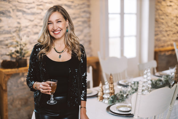 Dayna stands near a table that is set for the holidays. She holds a wine glass and smiles.