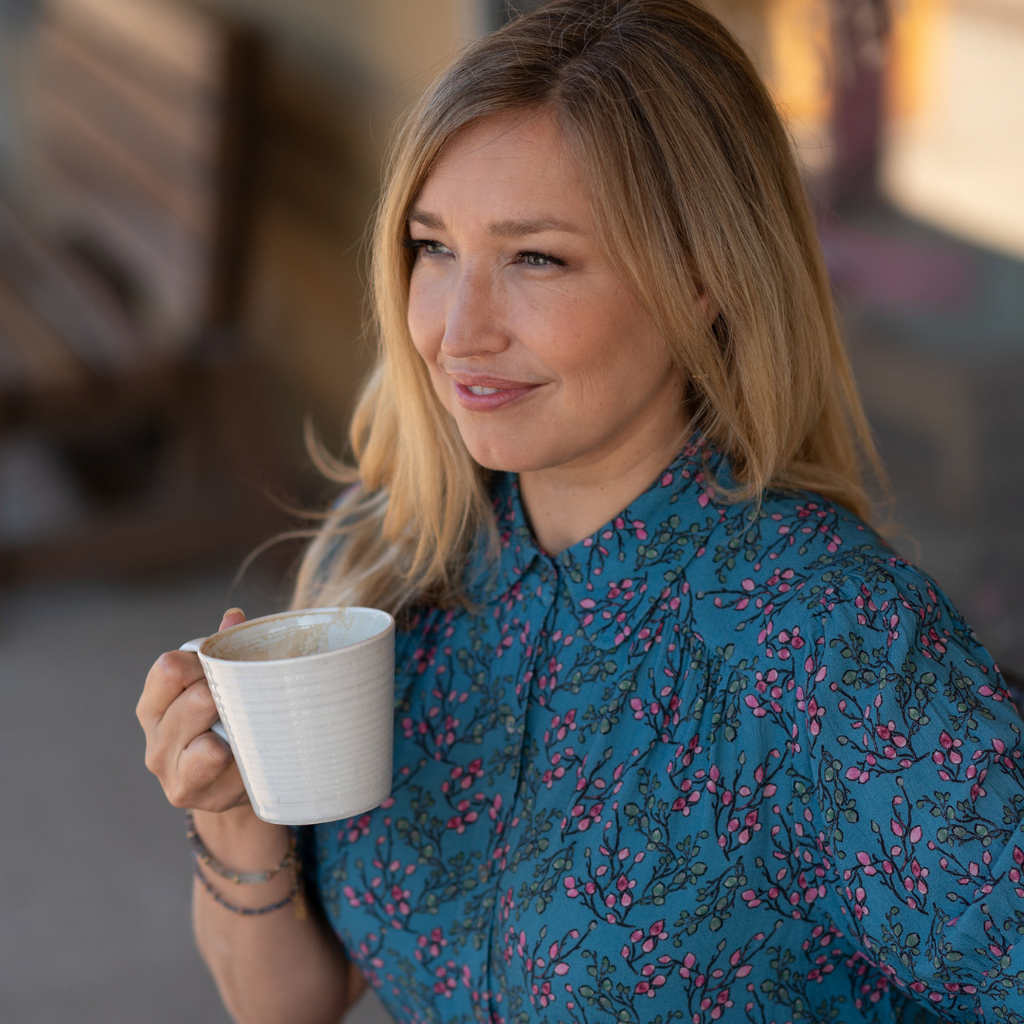 Dayna wears a blue blouse with a small floral print. She holds a coffee mug and looks into the distance.
