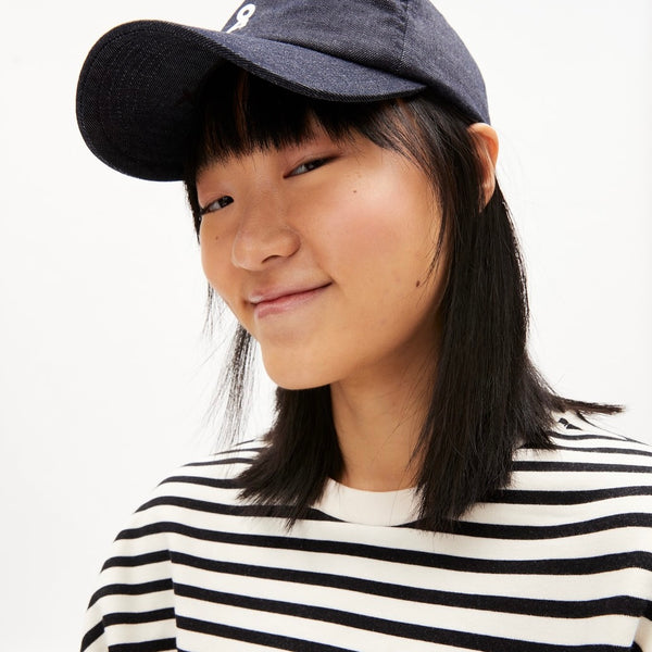 A woman is shown wearing a white T-shirt with black stripes. It is a sustainably produced T-shirt by Armed Angels. The model wears a baseball cap and smiles at the camera.