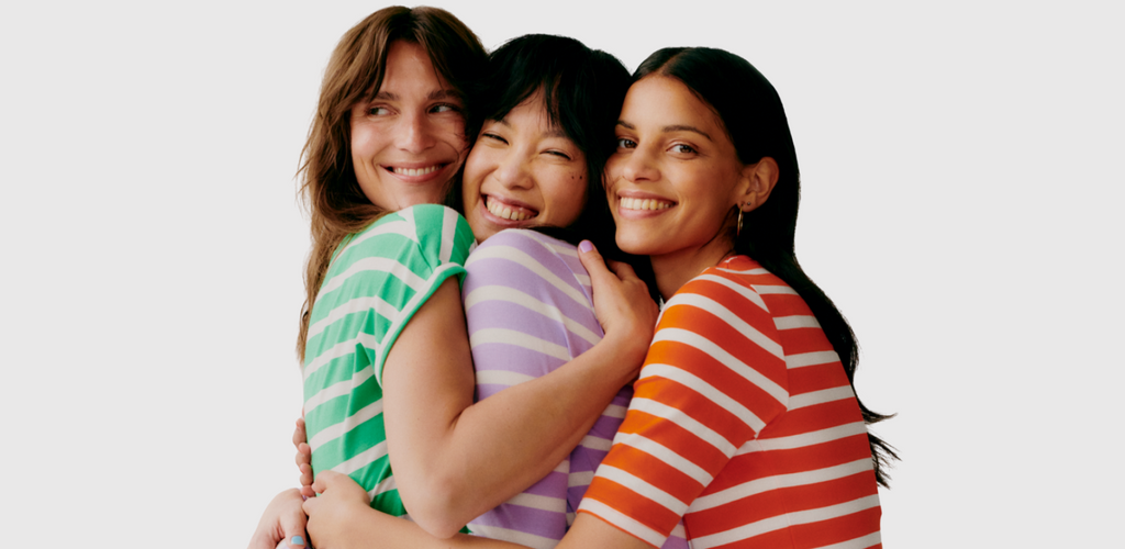 Three women are shown in brightly coloured T-shirts by B. Young. They hug and smile at the camera.
