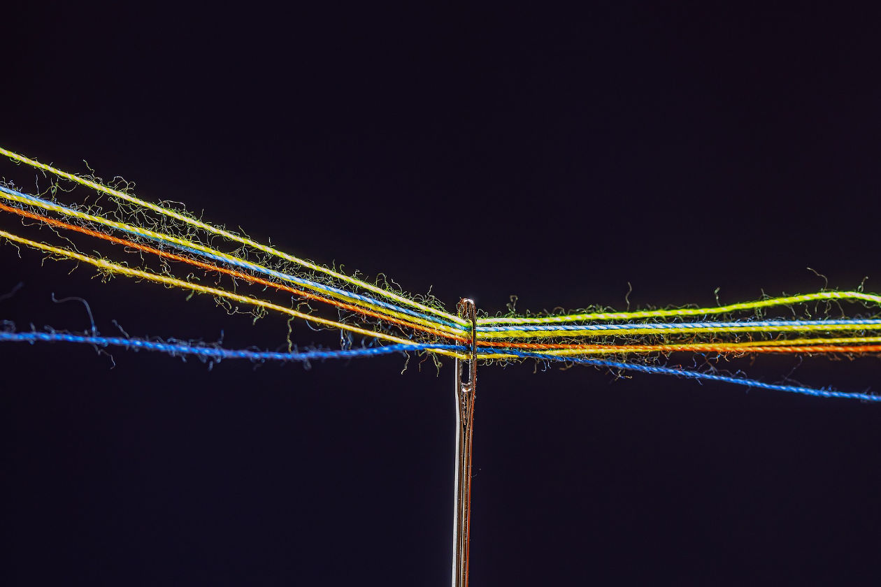 A close-up shot of a needle eye with several strands of thread inserted on it in different colors.