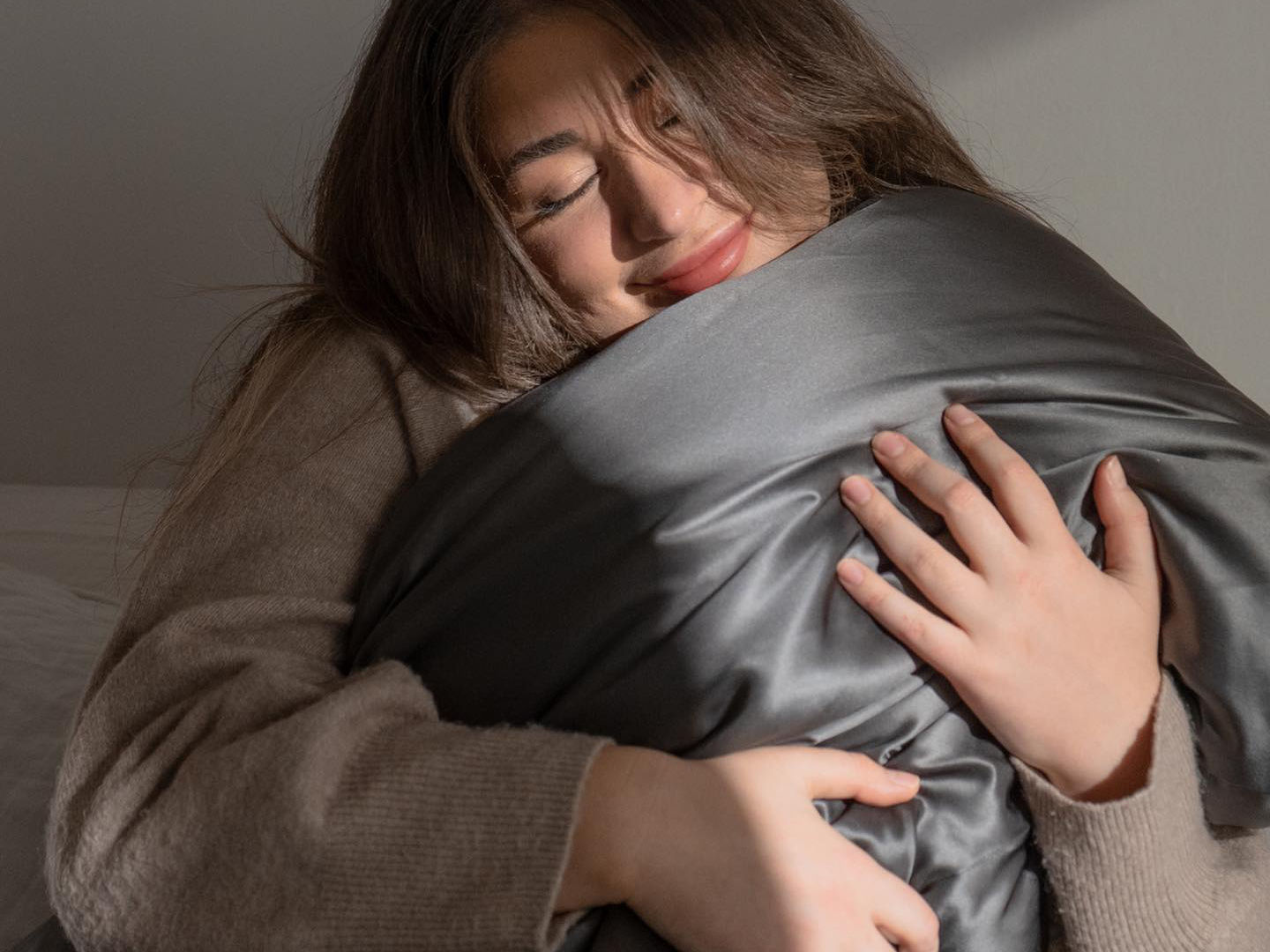 A smiling woman hugging a soft pillow covered with a Hush Silk Pillowcase in charcoal color.