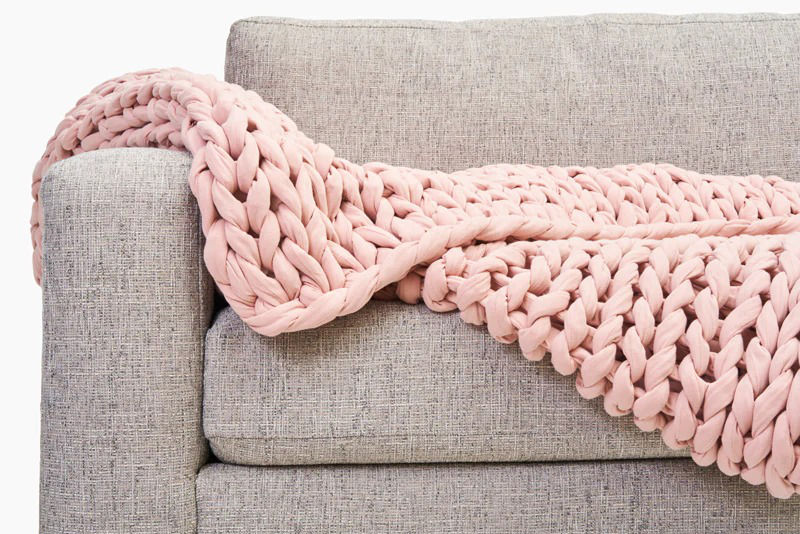 A Bearaby Cotton Napper in Evening Rose color draped over a grey sofa.