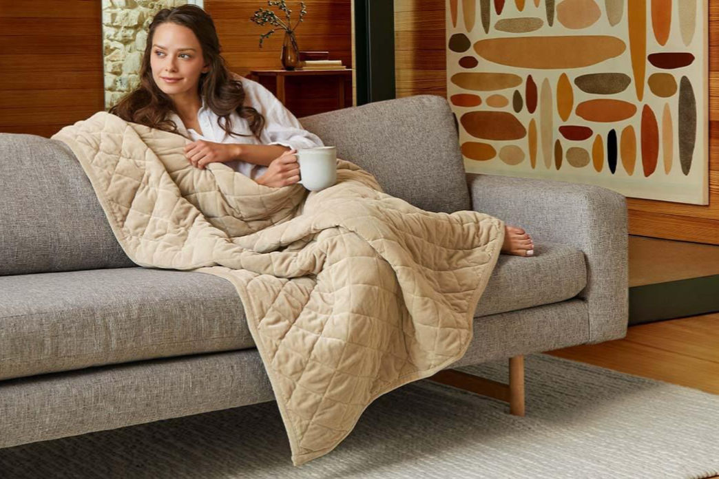 A woman sitting comfortably on a grey couch holding a cup while draped with a taupe Saatva Organic Weighted Blanket.