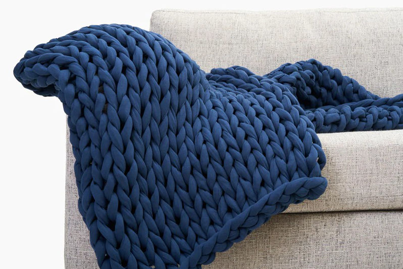 A Bearaby Cotton Napper in midnight blue color casually draped over a grey sofa.